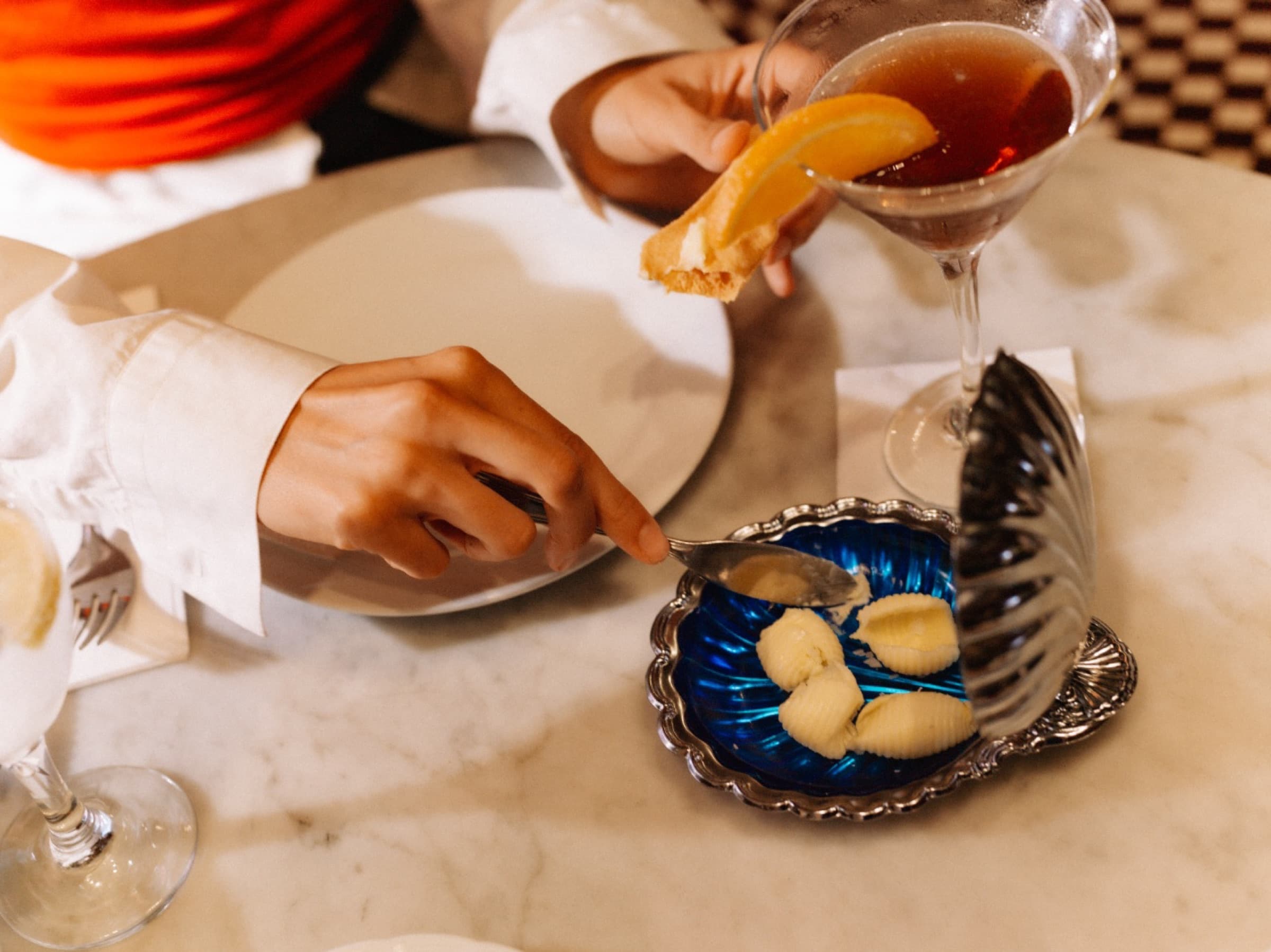 Een gast pakt een hapje van een blauw schaaltje naast een cocktail met sinaasappel